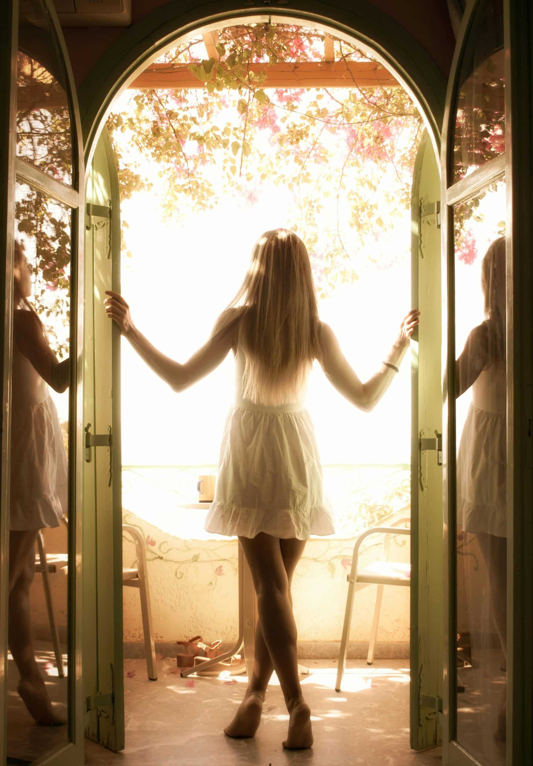 A young woman in a white dress enjoys morning sunlight on a balcony, surrounded by greenery.